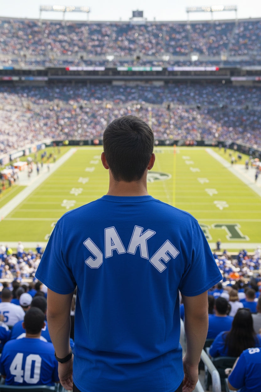 A royal blue short-sleeve t-shirt hanging on a black plastic hanger, showing the back with the name 'JAKE' printed in white arched block letters. The shirt is photographed in a home setting against a white door frame and brick wall.