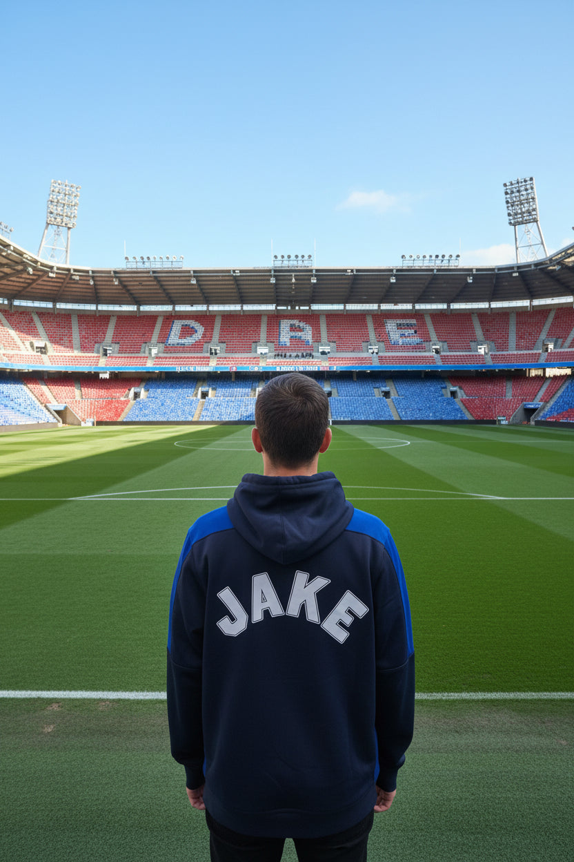 A navy blue and blue hooded sweatshirt shown from the back, featuring the name 'JAKE' printed in large white varsity-style letters with a dark outline. The hoodie is hanging in front of a doorway.