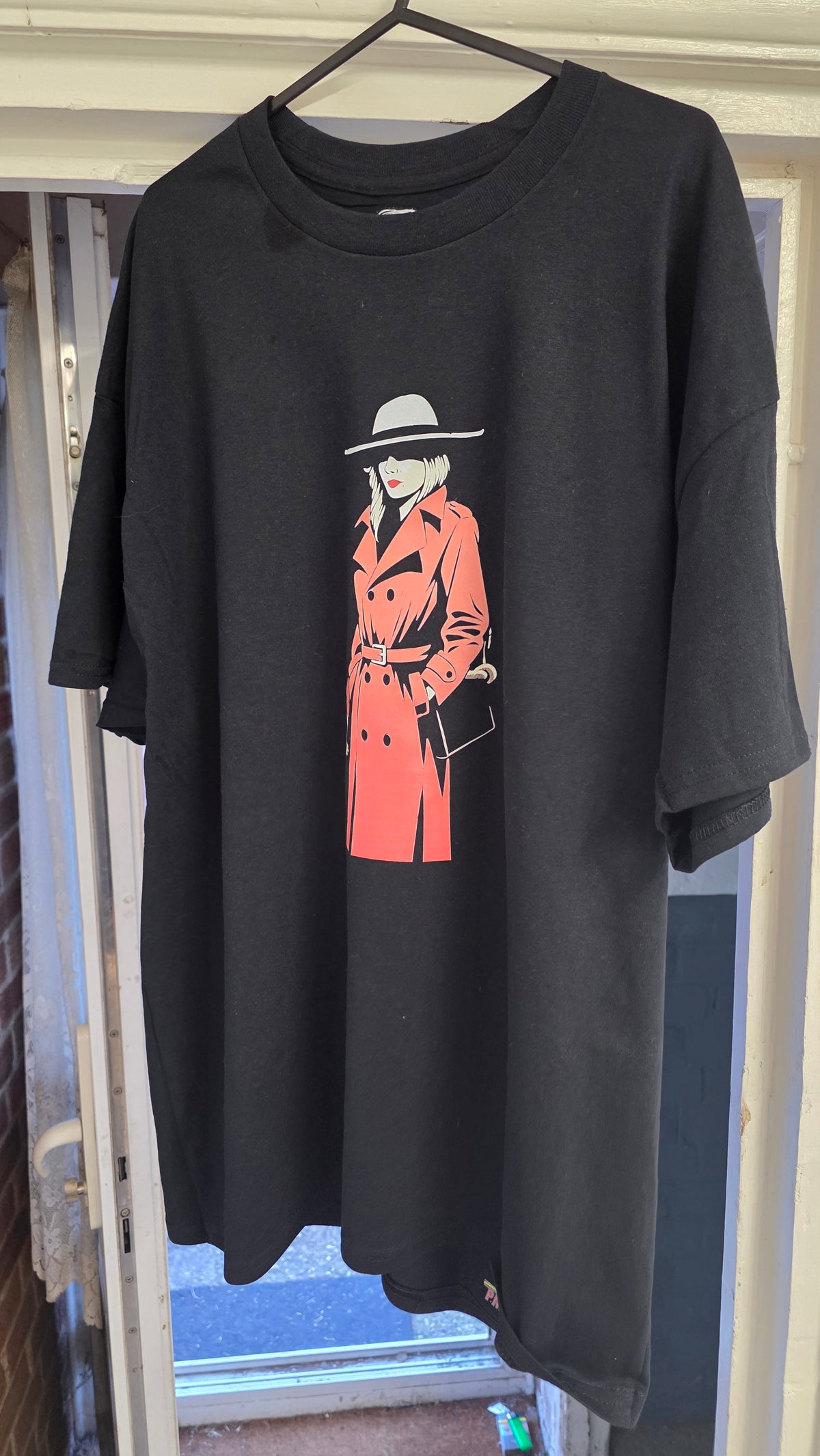 A black oversized short-sleeve t-shirt hanging on a hanger in front of a doorway. The shirt features a graphic print of a woman in a red trench coat and a gray hat.